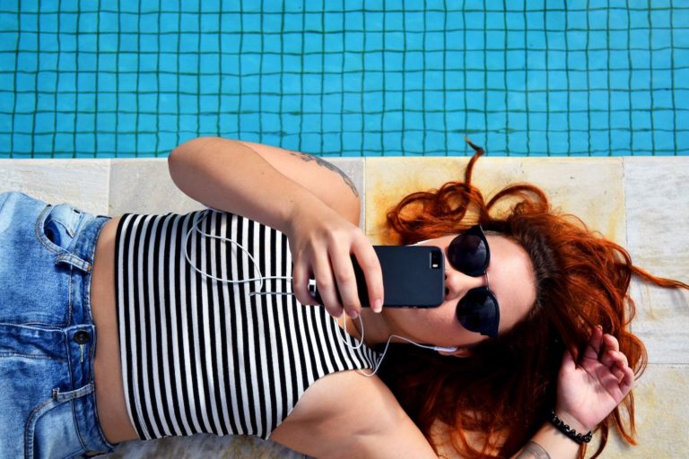 A young woman lies next to a pool, looking at her smartphone
