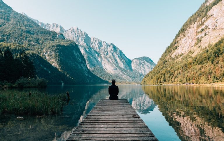 Man sits alone at the end of pier on a secluded lake.