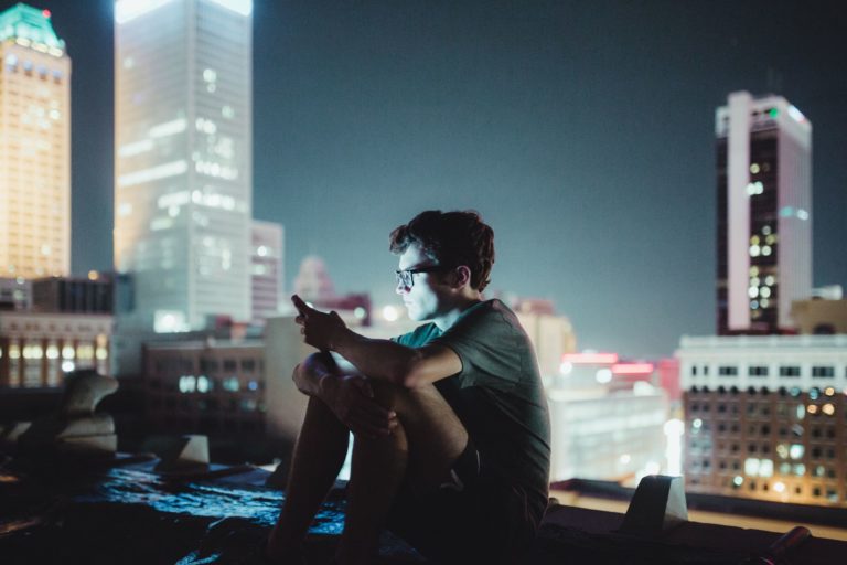 A young man seated on a rootop at night looks at a glowing smartphone