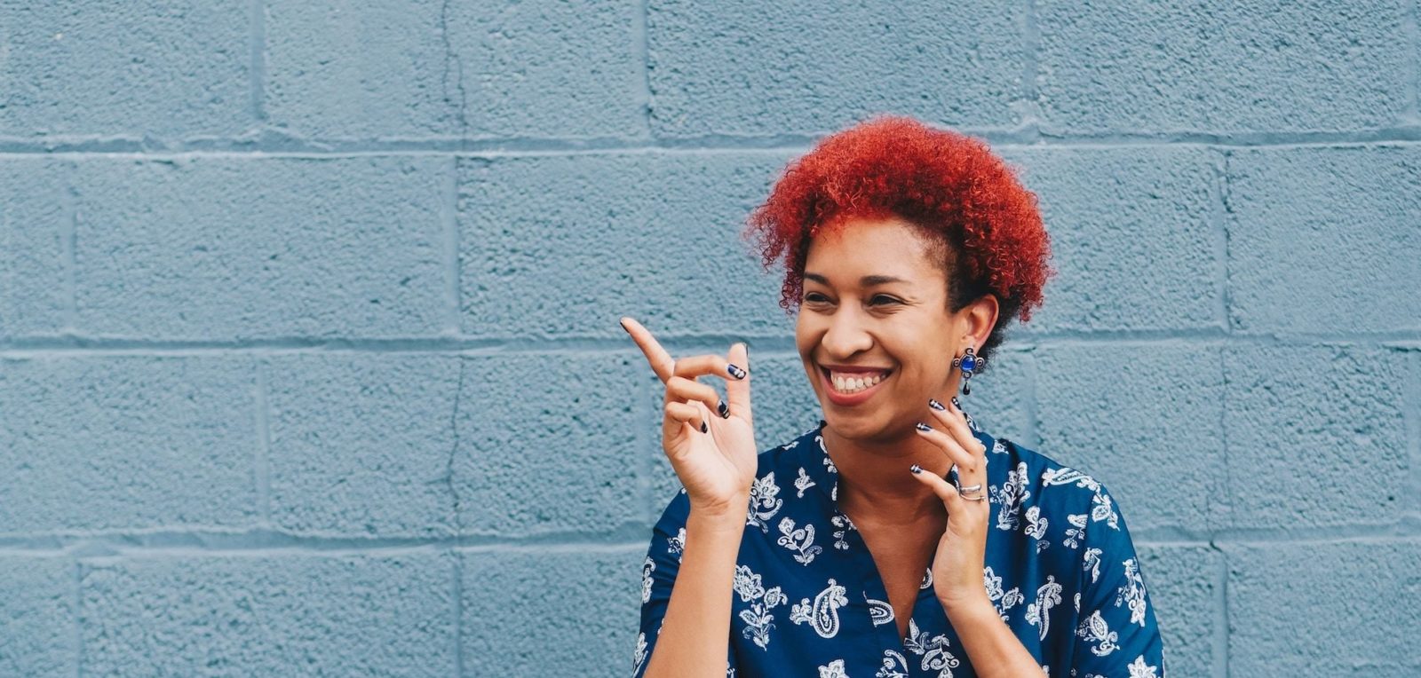 A woman with a bright red curls smiles and points in front of a blue wall