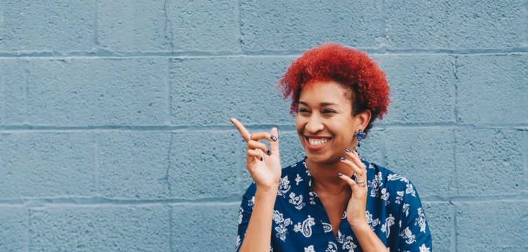 A woman with a bright red curls smiles and points in front of a blue wall
