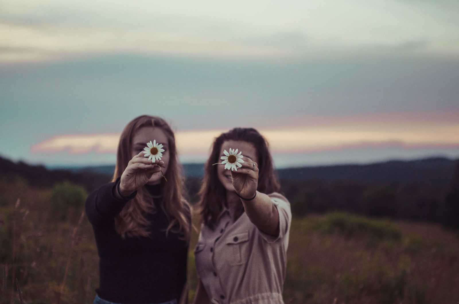 Two women in a field hold flowers up in front of their faces.