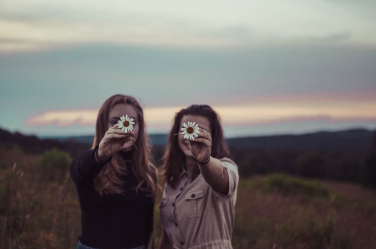 Two women in a field hold flowers up in front of their faces.