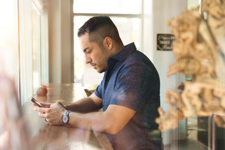 A man in a short-sleeved, button-up shirt reads an email marketing campaign in a cafe.