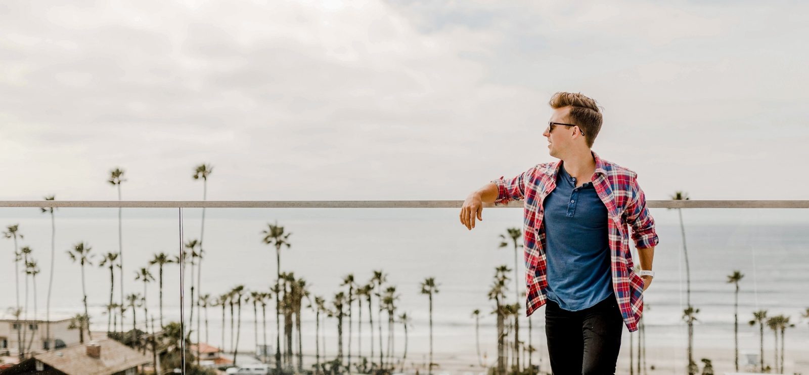 A man in a plaid shirt stands in front of a beach with palm trees.