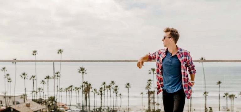 A man in a plaid shirt stands in front of a beach with palm trees.