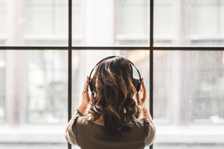 A woman listens to large headphones while looking out a glass window.