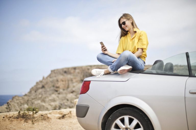 A woman wearing a yellow sweater browses a mobile app atop a car in the desert.