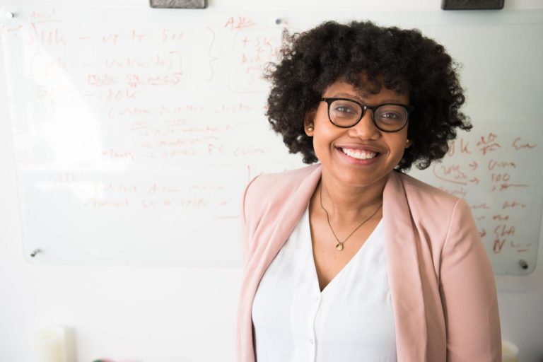 A woman wearing a blazer, button-up shirt, and glasses smiles while standing in front of a whiteboard.