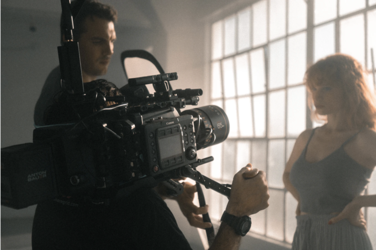 Two marketers use a digital video camera to record a young woman in front of a panel of glass windows.