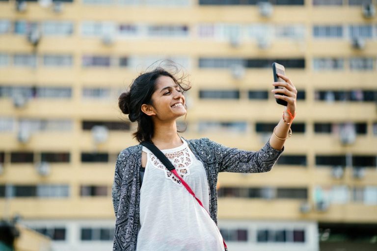 A smiling woman holds a smartphone in the air, gazing directly at it.