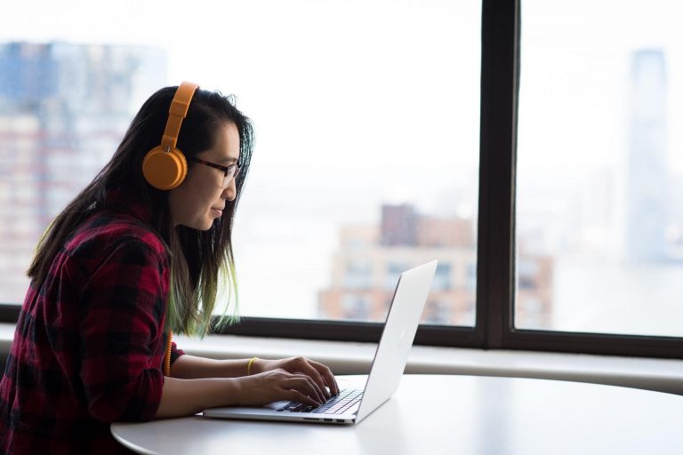 A woman wearing over-ear headphones publishes blog content using a laptop.