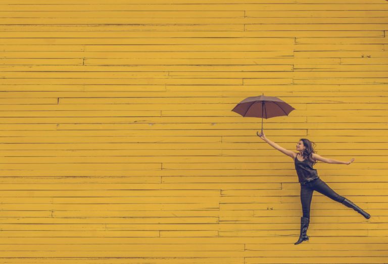 a woman floats through the air holding an umbrella with a brick wall behind her