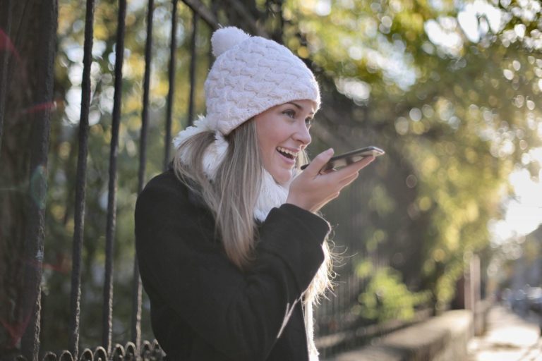 A woman wearing a winter hat uses a smartphone to conduct a voice search while standing on the street.
