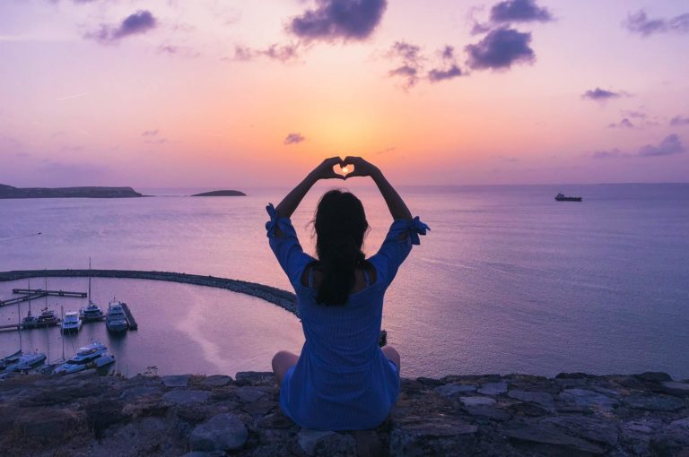 A woman sits on a rock and creates a heart symbol with her hands while watching the sun set.