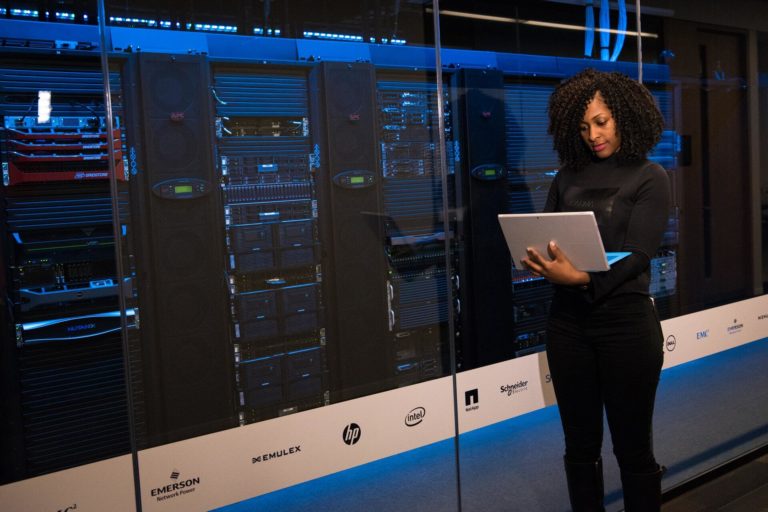 An expert content creator writes an article on a laptop while standing in front of technology stacks.