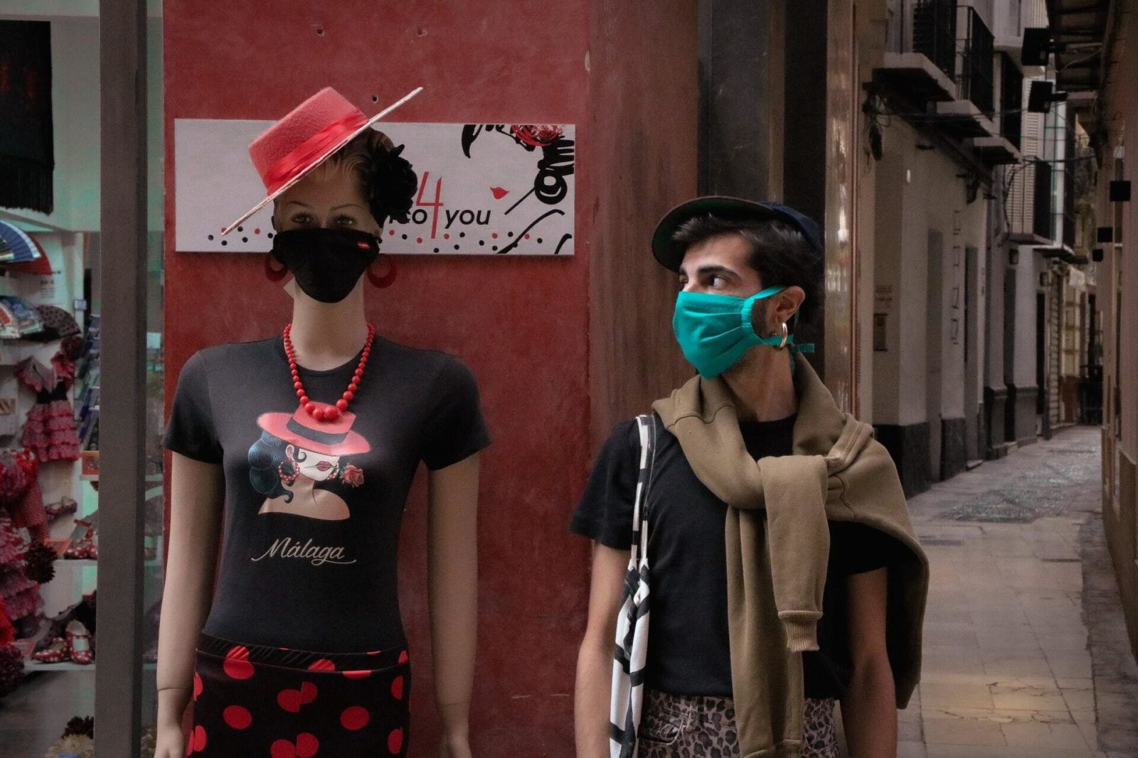 A masked shopper looks at a female mannequin that's wearing a mask and a hat.