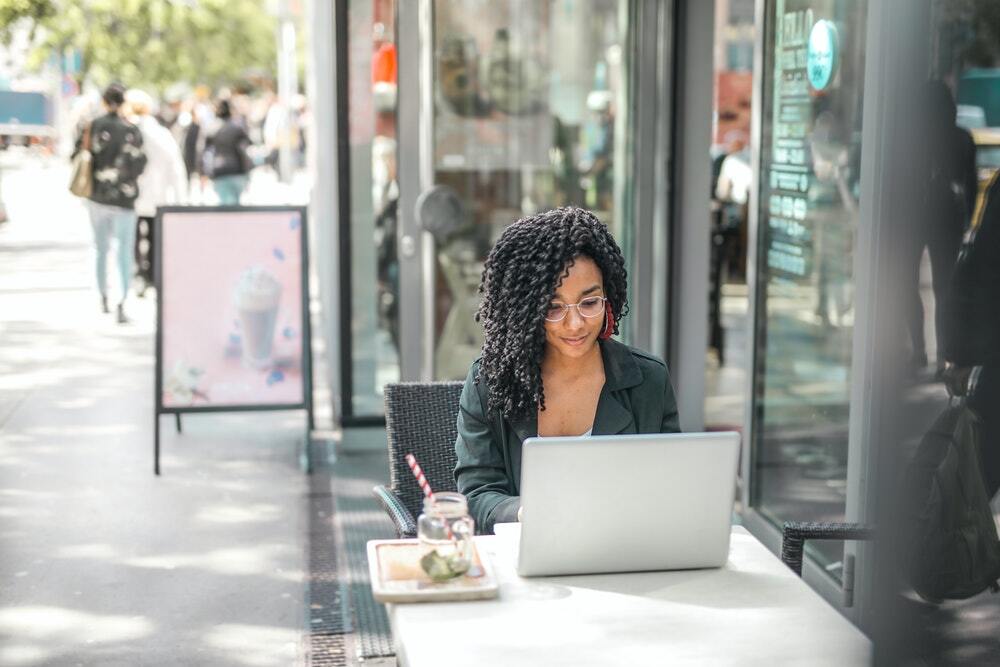 A young African-American freelance writer prices out a project on her laptop while seated outdoors at a cafe.