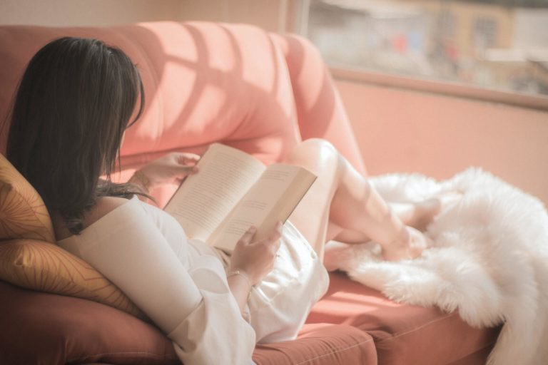 A woman with dark hair reads a book on a pink couch in a sunny room.