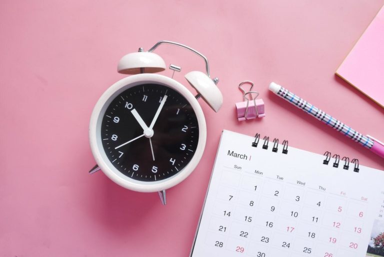 Light pink alarm clock, calendar, pen, and paper clip on deeper pink background.