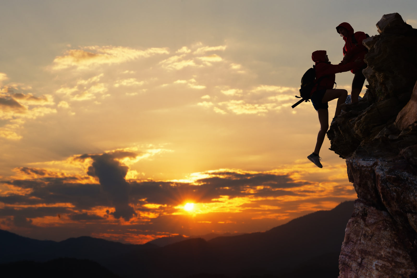 One hiker helping another up a cliff at sunset.