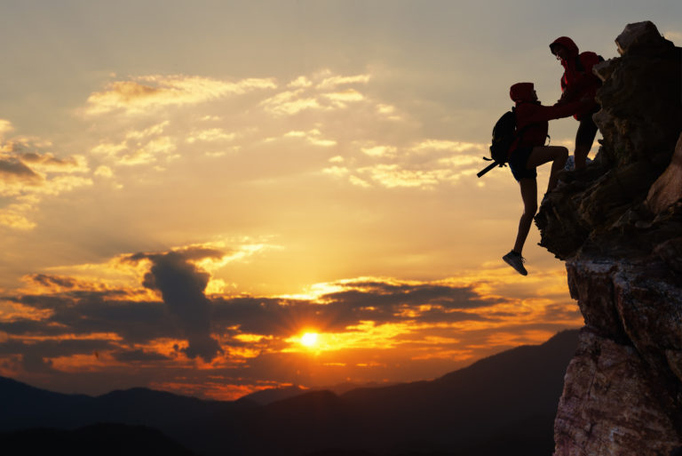 One hiker helping another up a cliff at sunset.