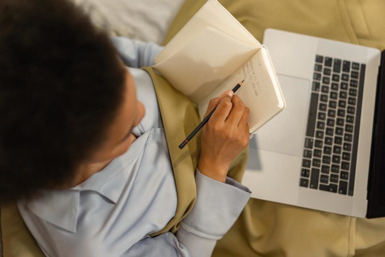 A person with laptop on lap and writing in notebook in hand.